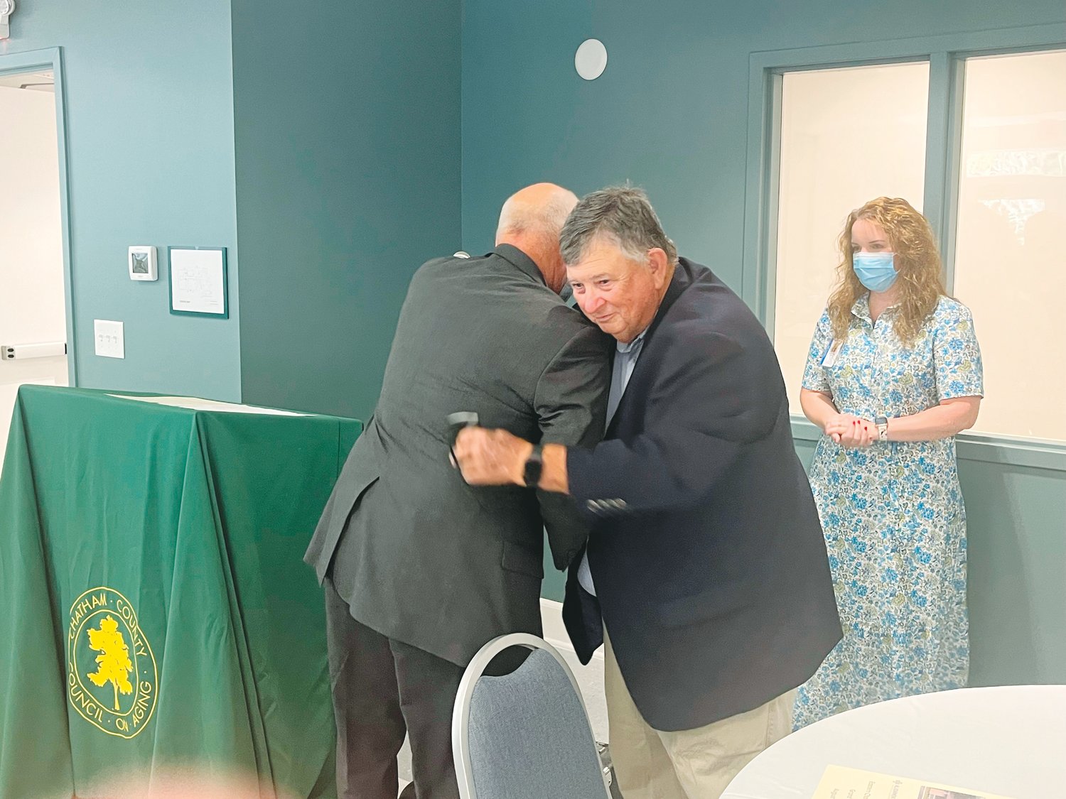 Johnny Shaw, president of the Chatham County Council on Aging board of directors, embraces retired Director Dennis Streets as current Director Ashlyn Martin looks on during the grand opening program of the renovated Eastern Chatham Senior Center last Friday in Pittsboro.